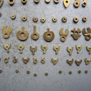 May include: An assortment of small, brass-colored watch parts, including gears, levers, and various shaped components. The parts are arranged on a gray surface, showcasing their intricate designs and varying sizes. The collection suggests a focus on precision and detail.