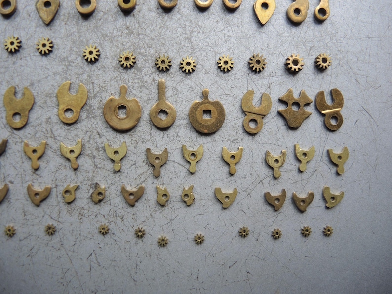 May include: An assortment of small, brass-colored watch parts, including gears, levers, and various shaped components. The parts are arranged on a gray surface, showcasing their intricate designs and varying sizes. The collection suggests a focus on precision and detail.