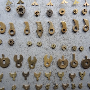 May include: An assortment of small, antique brass watch parts, including gears, levers, and other components, arranged on a gray surface. The parts vary in shape and size, showcasing intricate details and a vintage aesthetic.