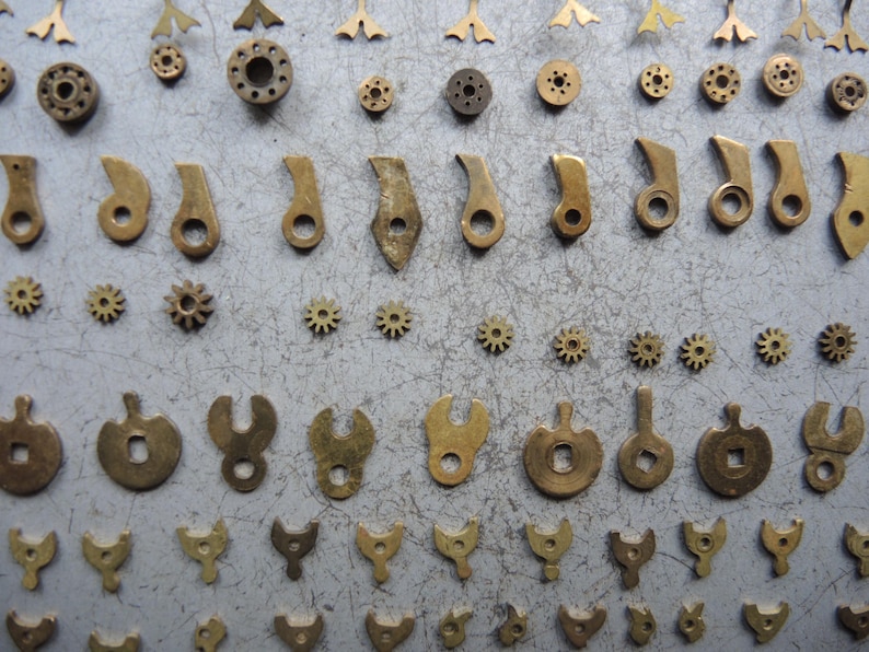May include: An assortment of small, antique brass watch parts, including gears, levers, and other components, arranged on a gray surface. The parts vary in shape and size, showcasing intricate details and a vintage aesthetic.