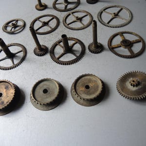 May include: An assortment of vintage clock gears and components, arranged on a gray surface. The gears vary in size and design, with a mix of circular and toothed elements. The metal has a weathered, aged appearance, suggesting antique origins.