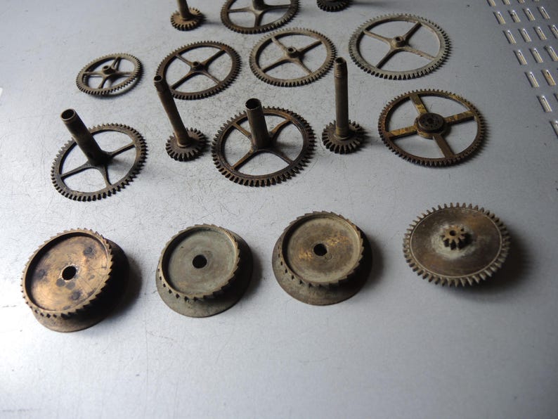 May include: An assortment of vintage clock gears and components, arranged on a gray surface. The gears vary in size and design, with a mix of circular and toothed elements. The metal has a weathered, aged appearance, suggesting antique origins.
