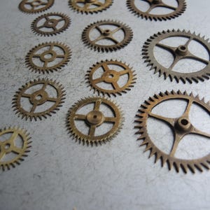 May include: Several small, brass-coloured gears of different sizes are arranged on a textured, grey surface. The gears feature detailed teeth and central holes, indicating they are parts of a mechanical device, such as a clock or watch mechanism.