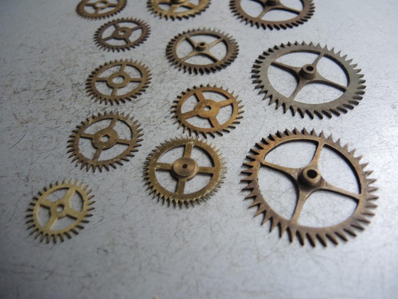 May include: Several small, brass-coloured gears of different sizes are arranged on a textured, grey surface. The gears feature detailed teeth and central holes, indicating they are parts of a mechanical device, such as a clock or watch mechanism.
