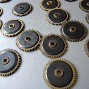 May include: A collection of small, circular gears with a brass center and toothed edges. The gears have a dark gray center and a gold-colored rim. The gears are scattered across a light gray surface.