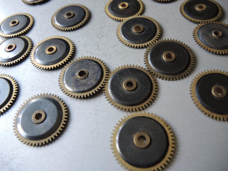 May include: A collection of small, circular gears with a brass center and toothed edges. The gears have a dark gray center and a gold-colored rim. The gears are scattered across a light gray surface.