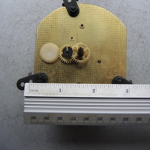 Op de afbeelding: Klokmechanisme met een messing plaat, tandwielen en een liniaal. De messing plaat heeft een getextureerd oppervlak en is ongeveer 10 cm lang. De tandwielen zijn klein en metaalachtig, met een wit plastic tandwiel zichtbaar.