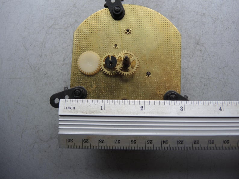 Op de afbeelding: Klokmechanisme met een messing plaat, tandwielen en een liniaal. De messing plaat heeft een getextureerd oppervlak en is ongeveer 10 cm lang. De tandwielen zijn klein en metaalachtig, met een wit plastic tandwiel zichtbaar.