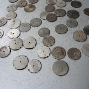 May include: A collection of small, circular metal gears with toothed edges and central holes. The gears vary in color from silver to a rusty brown, suggesting age or wear. They are scattered across a light-colored surface.