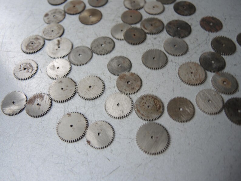 May include: A collection of small, circular metal gears with toothed edges and central holes. The gears vary in color from silver to a rusty brown, suggesting age or wear. They are scattered across a light-colored surface.