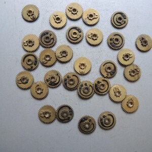 May include: A collection of small, brass-colored gears with intricate designs. Some gears have black detailing. The gears are scattered on a light gray surface, suggesting a mechanical or watchmaking theme.