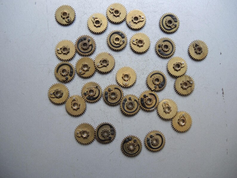 May include: A collection of small, brass-colored gears with intricate designs. Some gears have black detailing. The gears are scattered on a light gray surface, suggesting a mechanical or watchmaking theme.