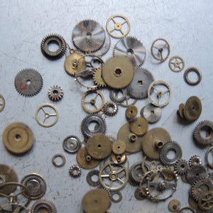 May include: A collection of various sizes of metal gears, in shades of gold, silver, and bronze, scattered on a light gray surface. The gears have intricate teeth and circular designs, suggesting a mechanical or industrial theme.