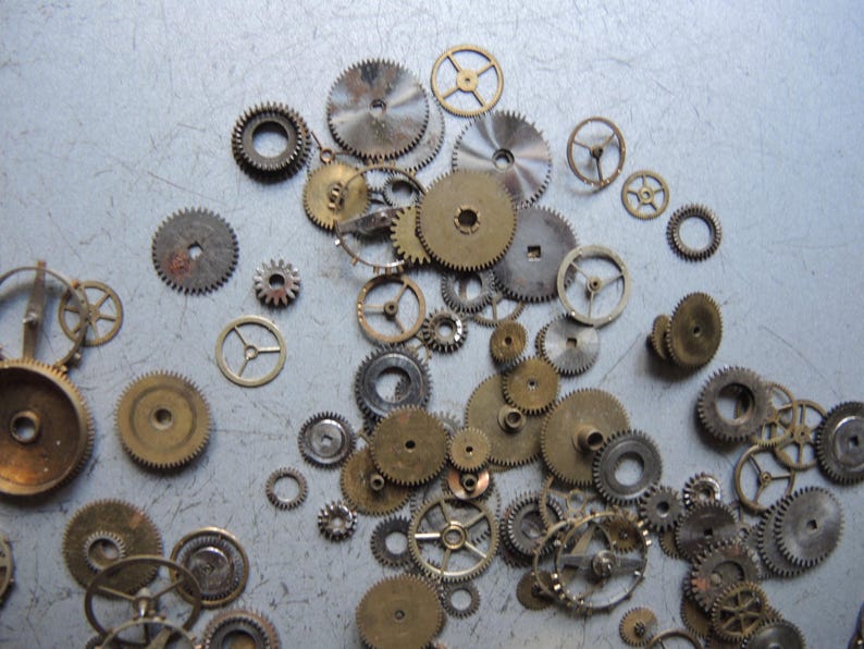 May include: A collection of various sizes of metal gears, in shades of gold, silver, and bronze, scattered on a light gray surface. The gears have intricate teeth and circular designs, suggesting a mechanical or industrial theme.