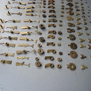 May include: A collection of small, vintage watch parts, including gears, levers, and balance wheels, in various sizes and shapes. The parts are made of brass and arranged on a gray surface, suggesting a repair or restoration project.