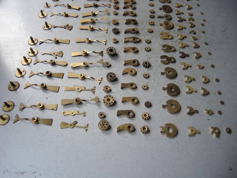 May include: A collection of small, vintage watch parts, including gears, levers, and balance wheels, in various sizes and shapes. The parts are made of brass and arranged on a gray surface, suggesting a repair or restoration project.