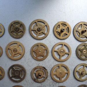 May include: A collection of various sized brass clock gears, arranged on a light gray surface. The gears have intricate teeth and central holes, showcasing their mechanical design. The gears are in different sizes and shapes.