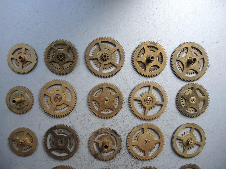 May include: A collection of various sized brass clock gears, arranged on a light gray surface. The gears have intricate teeth and central holes, showcasing their mechanical design. The gears are in different sizes and shapes.