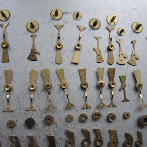 May include: A collection of vintage watch parts, including small brass gears, levers, and other components. The parts are arranged on a gray surface, showcasing their intricate designs and aged appearance. Ideal for watch repair or restoration projects.