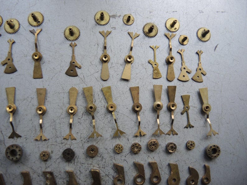 May include: A collection of vintage watch parts, including small brass gears, levers, and other components. The parts are arranged on a gray surface, showcasing their intricate designs and aged appearance. Ideal for watch repair or restoration projects.
