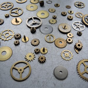 May include: A collection of various brass and silver-colored watch gears and cogs scattered on a gray surface. The gears vary in size and design, showcasing intricate details and mechanical components.