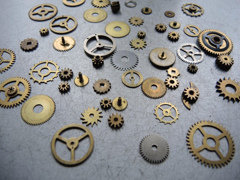 May include: A collection of various brass and silver-colored watch gears and cogs scattered on a gray surface. The gears vary in size and design, showcasing intricate details and mechanical components.