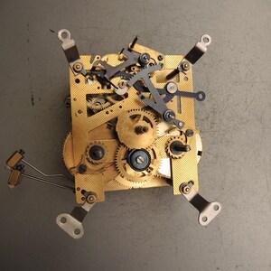 May include: Close-up of a clock mechanism with brass gears and metal components. The gears are intricately arranged on a gold-colored plate, with various levers and arms. The mechanism is set against a gray background, showcasing the detailed craftsmanship.