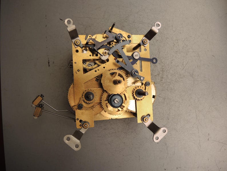 May include: Close-up of a clock mechanism with brass gears and metal components. The gears are intricately arranged on a gold-colored plate, with various levers and arms. The mechanism is set against a gray background, showcasing the detailed craftsmanship.