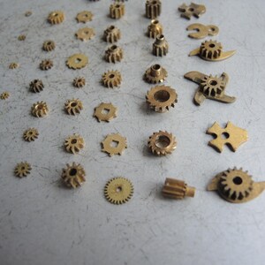 May include: An assortment of small, brass gears and mechanical components of varying sizes and shapes are scattered across a gray surface. The gears include circular, toothed, and uniquely shaped pieces, suggesting a collection of watch or clock parts.