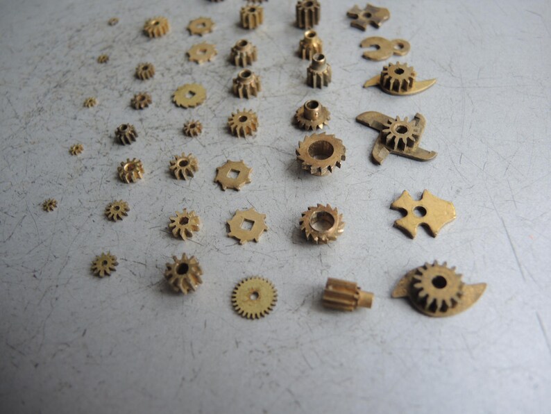 May include: An assortment of small, brass gears and mechanical components of varying sizes and shapes are scattered across a gray surface. The gears include circular, toothed, and uniquely shaped pieces, suggesting a collection of watch or clock parts.