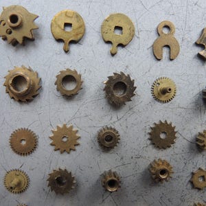 May include: A collection of small, antique brass clock parts, including gears, cogs, and other mechanical components. The parts vary in shape and size, with a warm, golden hue. These vintage pieces are arranged on a gray surface.