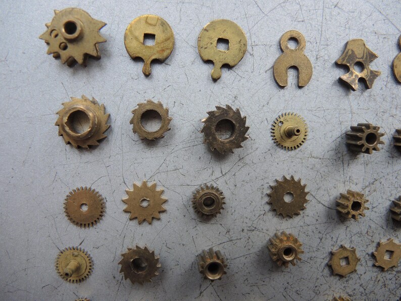 May include: A collection of small, antique brass clock parts, including gears, cogs, and other mechanical components. The parts vary in shape and size, with a warm, golden hue. These vintage pieces are arranged on a gray surface.