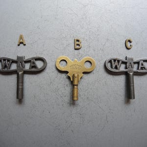 Puede incluir: Tres llaves de reloj antiguas y tres letras de latón, A, B y C, están dispuestas sobre una superficie gris. Dos llaves son plateadas con letras "WAA", y una es dorada con texto "WATERBURY CLOCK CO.".