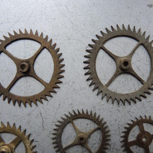 May include: A collection of small, bronze-coloured gears with pointed teeth. Each gear features a central hub with four spokes extending outwards. The gears are displayed on a light grey, metallic surface, evoking a mechanical or industrial aesthetic.