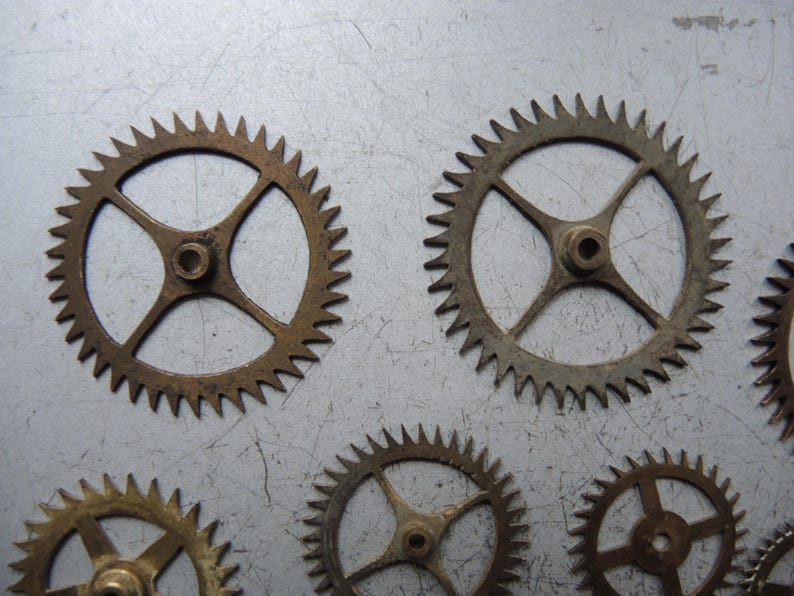 May include: A collection of small, bronze-coloured gears with pointed teeth. Each gear features a central hub with four spokes extending outwards. The gears are displayed on a light grey, metallic surface, evoking a mechanical or industrial aesthetic.
