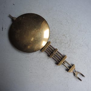 May include: A vintage brass clock pendulum with a circular weight and a rectangular suspension assembly. The pendulum has a warm, golden hue and is designed for timekeeping. It is a classic piece of horological hardware.