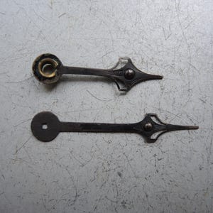 May include: Two antique clock hands. One hand has a circular end with a gold-coloured centre, while the other has a square hole. Both hands are dark metal with decorative, pointed ends and ornate detailing.