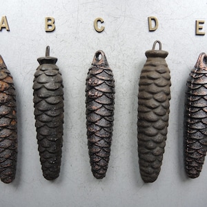 May include: Five antique, dark brown pine cone-shaped weights, each with a loop at the top for hanging. The weights are arranged in a row and labeled A through E with gold-colored letters. The cones have a textured, scale-like surface.