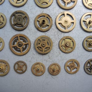 May include: A collection of various sized brass gears, arranged on a light gray surface. The gears have intricate teeth and central holes, showcasing their mechanical design. The gears vary in size and design, suggesting a variety of uses in clockwork or machinery.