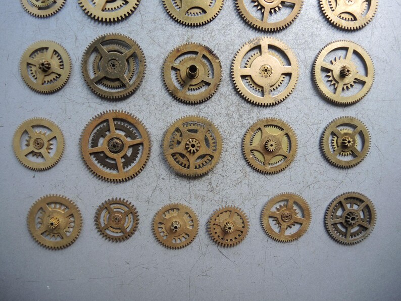 May include: A collection of various sized brass gears, arranged on a light gray surface. The gears have intricate teeth and central holes, showcasing their mechanical design. The gears vary in size and design, suggesting a variety of uses in clockwork or machinery.