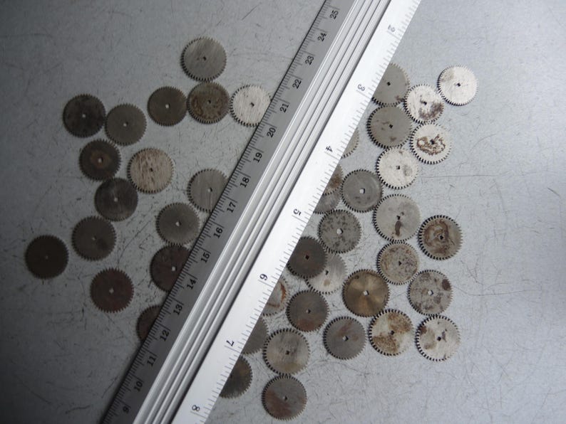 May include: A collection of small, circular metal gears with a ruler, likely for watchmaking or repair. The gears are various shades of gray and brown, with a ruler measuring up to 25 inches.