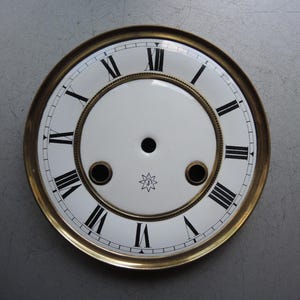 Könnte beinhalten: Ein Vintage-Zifferblatt mit weißem Hintergrund und schwarzen römischen Ziffern. Das Zifferblatt hat einen goldfarbenen Metallrahmen und ein kleines Sternmuster in der Nähe der Mitte. Zwei kleine Löcher sind in der Nähe der Mitte sichtbar.