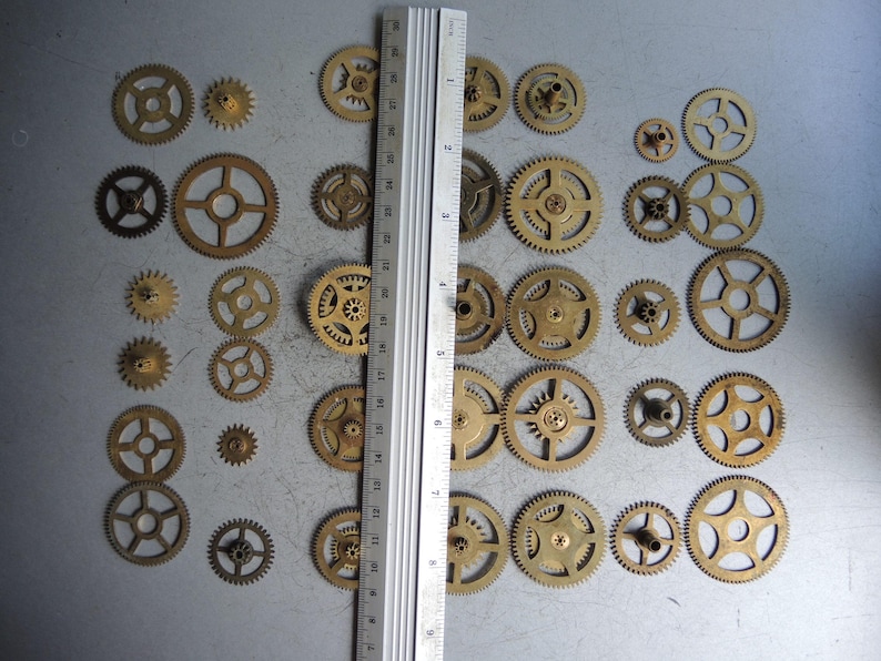 May include: A collection of small, brass-colored gears of various sizes, arranged on a gray surface. A metal ruler is placed vertically among the gears. The gears range from approximately 0.25 inches to 1.5 inches in diameter. The gears have different spoke designs.