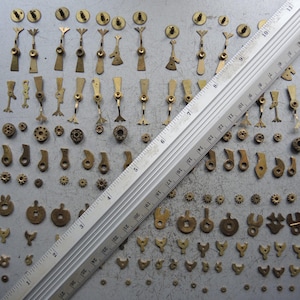 May include: A collection of small, antique brass watch parts, including gears, levers, and other components, arranged on a gray surface. A ruler is placed diagonally across the parts for scale. The parts vary in size and shape.