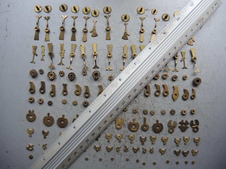 May include: A collection of small, antique brass watch parts, including gears, levers, and other components, arranged on a gray surface. A ruler is placed diagonally across the parts for scale. The parts vary in size and shape.
