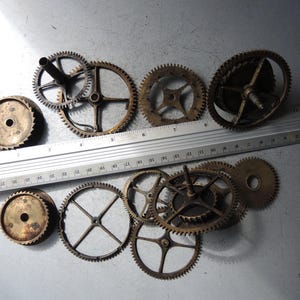 May include: A collection of vintage clock gears in various sizes and designs, with a metal ruler for scale. The gears are made of brass and show signs of age and wear. The ruler is marked in inches.