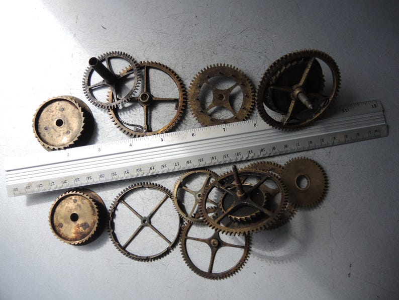 May include: A collection of vintage clock gears in various sizes and designs, with a metal ruler for scale. The gears are made of brass and show signs of age and wear. The ruler is marked in inches.
