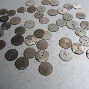 May include: A collection of small, circular metal gears with toothed edges and a central hole. The gears have a worn, aged appearance with varying shades of silver and brown, suggesting they are vintage or antique watch parts.