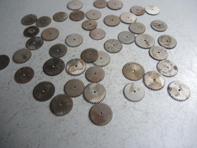 May include: A collection of small, circular metal gears with toothed edges and a central hole. The gears have a worn, aged appearance with varying shades of silver and brown, suggesting they are vintage or antique watch parts.
