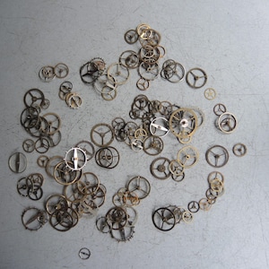 May include: A selection of small watch gears scattered on a grey surface. The gears vary in size and colour, with shades of gold, silver, and dark metal. Some gears have visible spokes and intricate designs, suggesting precision craftsmanship.